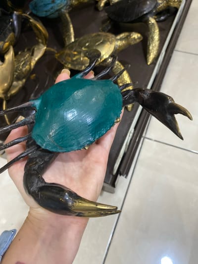A hand holds a green and black crab statue in front of a display of similar gold statues. The crab statue is black and has golden tips on its claws. The crab's body is a smooth green color. The person holding the crab statue has light skin and a visible blue denim jacket. The background is tile. It is daytime. This photograph was taken indoors, and it may be in a retail setting, likely a souvenir shop. Given the style of items shown, it is possible this image was taken in Nusa Dua, Indonesia, as the prompt states.