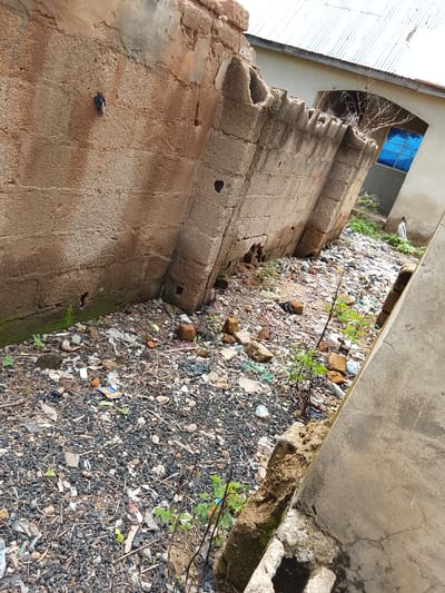 The media file depicts a scene of disrepair and neglect in a sun-drenched outdoor environment, likely in Sunkani, Nigeria. The primary subjects are the crumbling remains of a concrete structure, characterized by broken blocks and exposed rebar, suggesting a state of dilapidation or ongoing demolition. Adjacent to this, a wall constructed from rough-hewn, reddish-brown bricks stands, also showing signs of age and damage, with some sections missing, revealing the interior of the blocks.

The ground is covered in a thick layer of debris, consisting of small stones, gravel, and a significant amount of scattered refuse, including plastic bags, wrappers, and other discarded materials. Patches of moss and small, hardy green plants are growing amidst the rubble, indicating that nature is beginning to reclaim the space, despite the presence of human-generated waste.

In the background, a building with a corrugated metal roof and cream-colored walls is visible. A window with bright blue panes is a striking feature of this building. A lone, small figure, possibly a person, is standing in front of the building, adding a sense of scale and human presence to the otherwise desolate scene.

The lighting suggests it is daytime, with bright sunlight casting distinct shadows and illuminating the textures of the crumbling materials and the bright blue window. The overall atmosphere is one of abandonment and decay, juxtaposed with the resilience of plant life. There is no discernible text in the image. The location cues, such as the building style and the quality of light, align with the provided context of Sunkani, Nigeria.