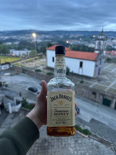 The image depicts a hand holding a bottle of Jack Daniel's Tennessee Honey whiskey against the backdrop of Vila Real, Portugal. The whiskey bottle is predominantly displayed in the foreground. The background features a cityscape with buildings, a church steeple, and a cloudy sky, likely taken from an elevated position. The focus is on the Jack Daniel's bottle, with the city serving as a secondary element to give the photo context. The hand holding the bottle appears to be wearing a green sleeve.