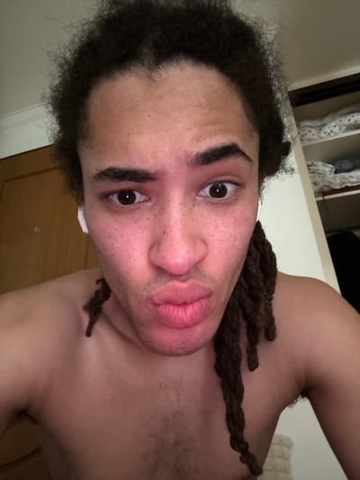 A close-up selfie of a light-skinned person with dark, curly hair and brown dreadlocks. The person has brown eyes, freckles, and full lips. They are shirtless and appear to be indoors. A partial view of a closet is visible in the background. There is no verifiable evidence of the location being Les Ulis, France.