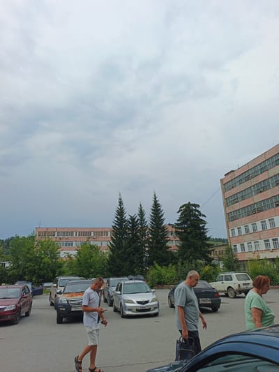 In Zlatoust, Russia, on an overcast day, several people are walking in a parking lot in front of two pink-toned buildings. In the foreground, a man in a white t-shirt and shorts walks to the right, looking at his phone. Behind him, another man in a grey t-shirt and dark pants walks to the right, carrying a shopping bag. A woman in a green shirt walks to the right in the middle ground. Several cars are parked in the lot, including a red sedan, a silver minivan, a dark SUV, a white SUV, and a black sedan. Evergreen trees are visible behind the cars and in front of the buildings.
