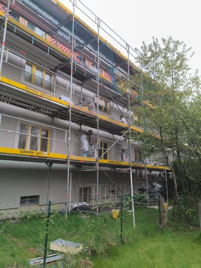 Here's a detailed description of the image:
**Scene Overview:**
The image depicts the exterior of a multi-story building undergoing renovation. The setting appears to be residential, with greenery suggesting a garden or yard.
**Location (Assuming Accuracy of Claim):**
The location is presumed to be in Münster, Germany.
**Subjects and People:**
Several construction workers are visible on scaffolding erected against the building. They are applying material to the exterior walls. The workers are wearing protective clothing.
**Objects:**
* **Scaffolding:** A substantial scaffolding structure is in place, spanning the height of the building. It is marked with the brand name "condor gerust."
* **Building:** The building has multiple windows, with yellow frames. Some windows are covered in plastic.
* **Construction Equipment:** Buckets and other construction implements are present on the scaffolding.
* **Fence:** A chain-link fence runs along the perimeter of the property, separating the building from the yard.
* **Vegetation:** Trees and grass are visible in the foreground, partially obscuring the lower part of the building and fence.
**Actions and Interactions:**
The workers are actively engaged in applying a coating (possibly plaster or paint) to the exterior walls of the building.
**Environment:**
The weather appears overcast. The environment is a mixture of construction and residential elements, indicating a renovation in a lived-in area.