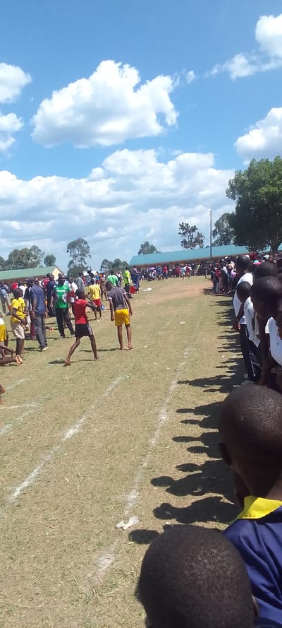 The photo shows a gathering of people on a grassy field, likely a sports event. The participants are primarily young men and boys, some wearing sports uniforms, others casual clothing. Several individuals are engaged in a race, running along white lines marked on the grass.  The audience is lined up along the edge of the field, mostly young men and boys, some wearing uniform shirts. Behind the field, there are buildings with a blue roof. The sky is blue with white clouds, and there are trees in the background.  The image was taken at a location with coordinates (latitude: 0.384, longitude: 34.801), which is near the town of Lubao in Kenya. There is a discarded plastic bag on the ground near the white lines.  The image was taken in a public area during daytime with a sunny sky.