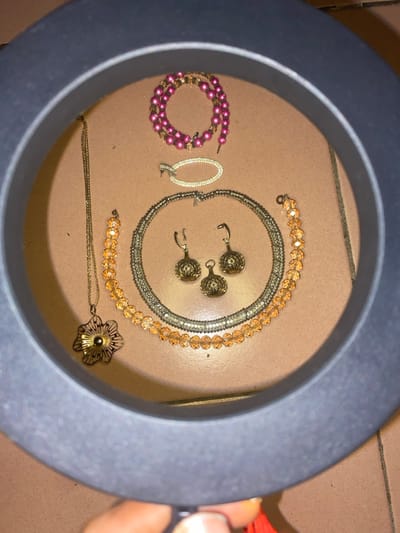 The image depicts a collection of jewelry items displayed within the dark gray circular frame of a ring light, held by a human hand, on a light reddish-brown ceramic tiled floor with visible grout lines. The scene is located in Ngleshi Amanfro, Ghana.
**Objects Present:**
* A coiled strand of approximately 70-80 spherical pink beads, approximately 5mm in diameter, interspaced with smaller clear beads or metal findings.
* An oblong, gold-toned or silver-toned metal brooch or clasp, approximately 4cm in length, encrusted with numerous small clear faceted stones along its surface.
* A gold-toned or silver-toned multi-strand flexible choker-style necklace, approximately 40cm in length, featuring a central segment adorned with multiple rows of small clear stones.
* A strand of approximately 25-30 faceted orange crystal beads, approximately 8mm in diameter, forming a necklace or bracelet.
* A thin gold-toned link chain necklace, approximately 45-50cm in length, with a central filigree flower-shaped pendant, approximately 4cm in diameter, featuring a radiating pattern.
* Two matching gold-toned drop earrings, each consisting of an ear hook and a hollow, spherical, textured or openwork filigree design element, approximately 2.5cm in diameter.
* One similar gold-toned, hollow, spherical, textured or openwork filigree design element, approximately 2cm in diameter, positioned separately from the earrings.
**Actions/Interactions:**
A human hand, with a light skin tone, is partially visible at the bottom center of the frame, holding the dark gray circular ring light. The jewelry items are laid out for display or photography.
**Environment:**
The items rest on a tiled floor, suggesting an indoor setting. The lighting appears artificial, originating from the ring light.