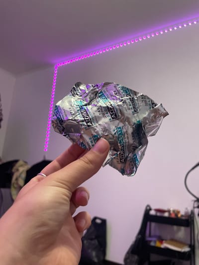 A crumpled silver foil blister pack, likely containing medication, is held in a person's left hand, which has a silver ring on the ring finger. The blister pack has text in a blue and white font. The background is a room with a white wall. A string of purple LED lights is mounted along the top of the wall, creating a border. To the right, a black shelf unit with various items is visible. The image is taken in a bedroom. The location of Vaganovo, Russia is irrelevant to the image content.