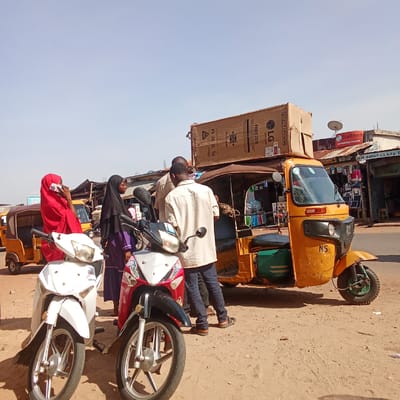The image depicts a bustling street scene in Lafia, Nigeria, characterized by a vibrant mix of people, vehicles, and commercial establishments.
In the foreground, two motorcycles stand parked on a dirt patch. To the left is a white motorcycle, and to the right, a red and black one. Three individuals are clustered nearby: a woman in a red hijab is on the phone, standing next to another woman in a black hijab and purple dress. A man in a light brown shirt and blue jeans appears to be in conversation with someone who is mostly out of frame.
Behind the group, a yellow "keke" or auto-rickshaw is prominently positioned. A large cardboard box, labeled "LG FREEZER," is strapped to the roof of the keke. The environment surrounding the keke is cluttered with goods and merchandise, indicative of a commercial area.
In the background, various shops and businesses line the street. A sign for "StarTimes AUTHORISED DEALER" is visible, along with other storefronts, suggesting a busy commercial zone. The sky is bright and clear, indicating daytime. The overall scene suggests a typical day in a bustling urban area of Lafia, Nigeria, with people engaging in commerce and transportation activities.
