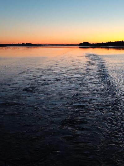 This image captures a wide-angle, static view of a large body of water at twilight in Khoroshevo, Russia. The sky transitions horizontally from light blue overhead to a deep orange and yellow at the horizon line. A continuous, dark silhouette of forested land or low hills delineates the distant horizon. The water surface exhibits dark blue-gray tones with numerous ripples and wave patterns across the foreground and mid-ground. A prominent wake-like disturbance or current is visible on the water, extending from the mid-left towards the mid-right. The water surface near the horizon reflects the sky's warm orange light. Faint mist rises from the water surface adjacent to the distant land on the left. A single green marker buoy is situated in the mid-ground water, to the left of a distinct land protrusion on the horizon. There are no individuals, animals, or active vessels present within the frame.