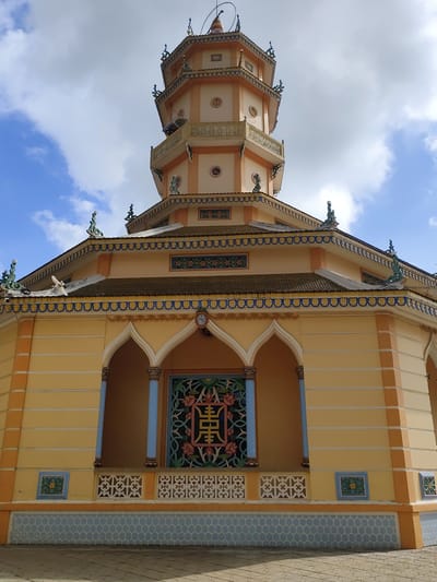 The image shows a low-angle shot of a yellow, multi-tiered pagoda with ornate architectural details. The pagoda has a pointed roof with decorative figurines and intricate trim along its edges. The facade features arched openings with colorful latticework and columns. A large, square window with a central Chinese character in a decorative frame is visible. The building is set against a backdrop of a bright blue sky with scattered white clouds. The ground in front of the pagoda is paved with square tiles. The pagoda appears to be located in a town or city, possibly a place of worship, given its design and scale. It is likely the Minh Tinh Pagoda in Lộc Quý, Vietnam, known for its distinct architecture.
