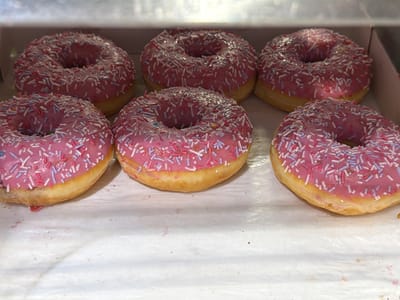 In Podolsk, Russia, a box of six pink frosted donuts with white, pink, and blue sprinkles is displayed. The donuts are arranged in two rows, three in the front and three in the back, on a white paper lining inside a cardboard box. The lighting suggests an indoor setting, possibly a bakery or store.