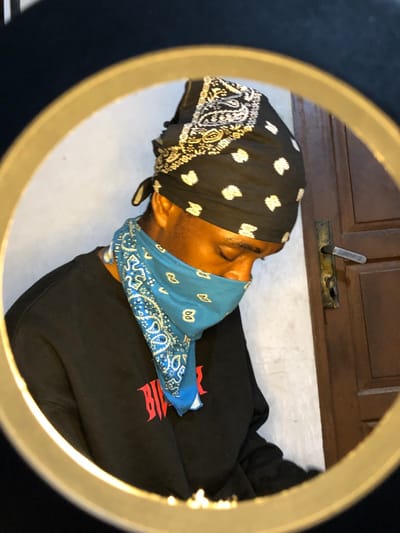 A person wears a black bandana with white patterns on their head, and a blue bandana with white paisley patterns covering their face. They are wearing a black sweatshirt with a red design on the chest. The person is centered in the frame of a ring light, with a door and light-colored floor visible in the background.