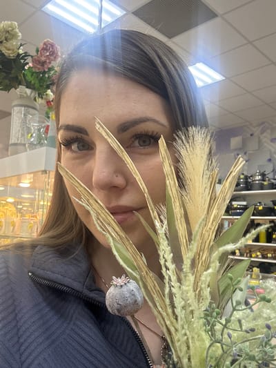 A young woman with brown hair and green eyes is facing the camera, partially obscured by decorative floral arrangements. Her hair is styled with darker roots transitioning to lighter ends. She has a nose piercing and is wearing a dark blue textured zippered jacket. She is indoors. Behind her is a display case with assorted items and rows of shelves with kitchenware, suggesting a retail environment. A glimpse of artificial flowers in a vase is seen in the lower left. The overall setting seems to be a shop, possibly in the city of Klin, Russia, based on additional, unavailable context.