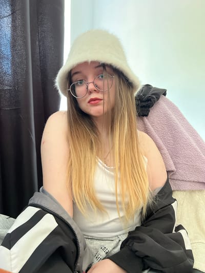 A young woman is lying on a black surface, likely a sofa or bed, wearing a white tank top, a furry white hat, and round glasses. She has long blonde hair and fair skin. She is partially covered by a black and white jacket. A lavender blanket is behind her. The location is not discernible from the image. Pushchino, Russia is not discernible from the image.
