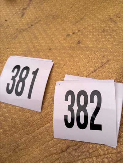 The image shows two white stickers with the numbers "381" and "382" printed in black. The stickers are placed on a textured yellow surface. The location is not discernible.