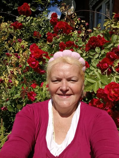 A woman with blonde hair, wearing a pink headband, a magenta cardigan over a white top, and a yellow necklace, smiles at the camera. She is positioned in front of a dense bush of red roses in full bloom, with green foliage. Behind the roses, a portion of a dark brown wooden house with a white-trimmed window is visible. The scene appears to be outdoors on a sunny day. The location is identified as the city of Krasnyy Mayak, Russia.