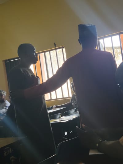 The image captures an indoor scene within a classroom or meeting room in Keffi, Nigeria, strongly backlit by natural light streaming through multiple barred windows on the right. The walls are a pale yellow/beige.
In the center, two adult males stand engaged in interaction. The figure on the left is seen in profile, facing right, wearing a dark, long-sleeved garment, appearing to speak or present. His right hand is partially raised towards his chest. The figure on the right is seen from the back, wearing a dark, long-sleeved top and a traditional dark-colored cap; his left arm is extended forward, directed towards the standing figure on the left, suggesting a gesture or physical contact.
Several other individuals are present, seated at desks. On the far left, a man wearing a light-colored top and a patterned cap is partially visible, seated. In the foreground on the right, hands and forearms of seated individuals are visible on desks, alongside papers, indicating other participants or observers in a discussion or lesson. The overall scene suggests an active conversation or instruction session.