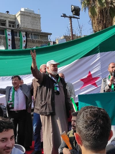The image captures an outdoor gathering in Al-Qanawat Municipality, Syria, under a clear blue sky. A large banner, approximately 10 meters wide, displaying the Syrian opposition flag (green top stripe, white middle stripe with three red stars, black bottom stripe) forms the background. Two smaller, vertically hung Syrian opposition flags are visible on a building in the upper left background.

In the central foreground, a middle-aged male with a grey beard and a white cap, wearing a dark brown jacket over a light brown tunic, raises his right hand in a V-sign. He has a dark lanyard around his neck. To his immediate right (viewer's left), a man in a grey suit jacket and white shirt, adorned with a Syrian opposition flag scarf, smiles broadly with both arms slightly raised. A black drum is partially visible next to him. To the central figure's immediate left (viewer's right), a bearded man in a grey jacket holds a microphone to his mouth, also wearing a Syrian opposition flag scarf.

Approximately five additional male figures are partially visible in the mid-ground, either behind or to the sides of the central figures. One man in the lower right foreground is seen from behind, obscuring part of the scene. Directly behind him, another male holds a rifle with a distinct orange-brown foregrip and stock. The environment includes a multi-story building with multiple windows in the upper left background and a utility pole topped with a large floodlight in the upper right, with sparse tree foliage and a palm tree visible behind it.