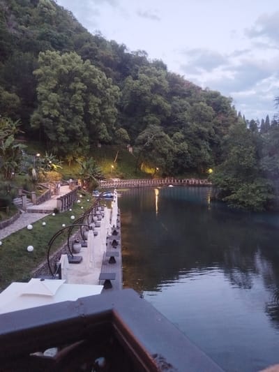 The image depicts a serene waterfront scene in New Athos, Abkhazia. A tranquil body of water, reflecting the overcast sky and lush green trees, dominates the right side of the frame. On the left, a paved walkway meanders alongside the water, lined with white umbrellas and outdoor seating, suggesting a restaurant or cafe area. Several large, white spherical lights are scattered across the grassy embankment. In the background, a dense forest covers a hillside, with lights visible in the distance, indicating structures or pathways within the foliage. A railing in the foreground suggests the vantage point is from a balcony or elevated platform. Two individuals are seen strolling on the walkway, engaged in conversation. The overall atmosphere is peaceful and inviting, hinting at a resort or recreational area.