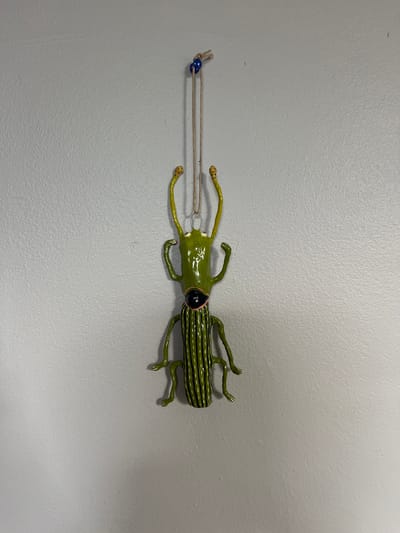 A painted metal sculpture of a green beetle is displayed horizontally on a textured, light gray wall. The beetle is oriented with its head to the left and its segmented abdomen to the right. The head is small and green, with black eyes. Its antennae are thin and yellow, and it has two thin, yellow arms that extend forward. The beetle's body is a bright green, and its abdomen is decorated with thin, dark green or black stripes. Four thin, green legs are visible, two on each side, extending downward from the body.
A thin, tan string is attached to the beetle's head. The string extends to the left, and a small, blue bead is threaded onto it. The string is taut, suggesting the beetle is hanging or suspended. The background is a plain, light gray wall with a slightly textured surface, typical of interior walls. The lighting appears to be artificial, casting a soft, even illumination on the scene. There is no indication of the time of day, weather, or specific location beyond the general context of being indoors. No text is visible in the image.