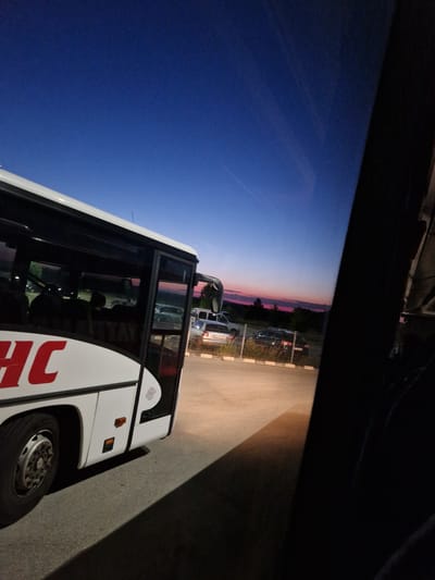 The image depicts a sunset scene in Bahovitsa, Bulgaria, as viewed from inside a bus. The sky transitions from a deep blue overhead to shades of pink and orange near the horizon, with the silhouette of trees visible against the colorful sky. In the foreground, the side of a white bus with red lettering is prominent. Through the bus windows, several cars and a white van are parked in an outdoor lot, separated by a fence. The ground is asphalt. The lighting suggests it is dusk, with artificial lights illuminating some areas and casting shadows. The perspective is angled, looking out of the bus towards the sunset and the parked vehicles.
