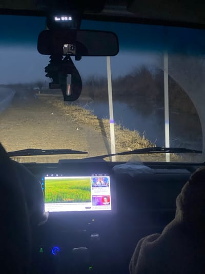 The scene depicts an interior view from a vehicle at dusk or early night in Batash, Uzbekistan. A road is visible to the left through the windshield, illuminated by the vehicle's headlights. To the right of the road, a body of water, possibly a canal or ditch, runs parallel, bordered by dry, light-colored vegetation. Two white vertical poles are partially visible near the water. The distant background is obscured by darkness or fog, revealing bare trees or shrubs.
Inside the vehicle, an overhead console displays "00 18:16." A rearview mirror, a dashcam, a pine-shaped air freshener, and a dark, elongated hanging ornament are attached near the top of the windshield. The windshield wipers are at rest. On the dashboard, a large digital infotainment screen displays a video of a green field on the left and various smaller video thumbnails on the right, with text indicating "Yil Sallama & Ibrox Boys - Bozi Sangli Haydar Mix 2024 (Yeni Hatira Songasi) 2024." A hand, likely belonging to the passenger, is seen interacting with this screen. The left arm and shoulder of a person (possibly the driver) are visible in a dark garment on the left. The right arm and shoulder of a passenger, wearing a brown, textured garment, are visible on the right. Below the infotainment screen, vehicle controls feature blue illumination. A crumpled white tissue or paper rests on the dashboard in front of the passenger.