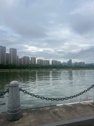 The image shows a waterfront view in Liandu District, China, on a cloudy day. In the foreground, a stone bollard with a thick metal chain attached to it runs along the edge of a paved walkway. Beyond the walkway, a body of water stretches towards the opposite shore, where a line of trees and several high-rise buildings are visible. Further in the distance, more buildings and mountains can be seen under the overcast sky. The water's surface is rippled, reflecting the muted light.