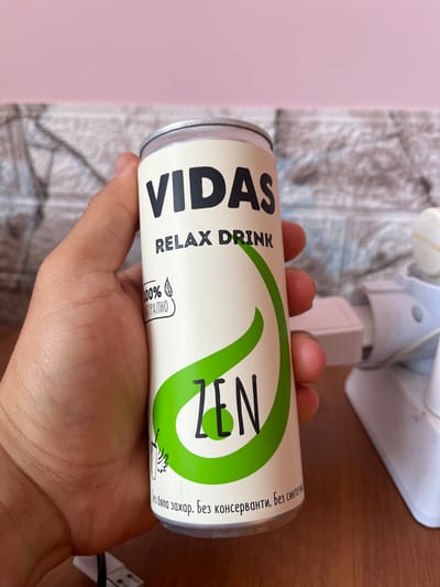 A hand holds a white can of "VIDAS RELAX DRINK ZEN". The can has black text and a green leaf-like design. Below the "ZEN" text, there is smaller text in Bulgarian stating "Без бяла захар. Без консерванти. Без синтетични". The background is blurred, showing a light pink wall and a grey textured surface, possibly a brick wall. There is also a white object to the right of the can, which appears to be a piece of electronic equipment. The image was taken in the city of Vratsa, Bulgaria.