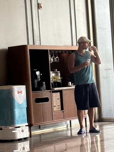 In the Huangpu District of China, a young man wearing a teal tank top, black shorts, and a white baseball cap stands to the right of a modern wooden cabinet. He holds a white cup to his lips with his right hand and a smartphone in his left. The cabinet features a black coffee machine, a glass dispenser filled with yellow items, and wine glass holders. To the left of the cabinet is a light blue and white cylindrical service robot with a red bow tie graphic. The scene takes place indoors with a tiled floor that reflects the objects above.