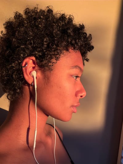 A person with dark curly hair and a pierced ear is lying down, looking upwards and towards the left side of the frame. They are wearing white earbuds, the cord of which runs across their neck and upper chest. The person's skin tone is reddish-brown, illuminated by a warm light. The background is a plain, light-colored wall, with a darker shadow near the top edge. The scene is indoors. There is no indication that the image was taken in Champs-sur-Marne, France.