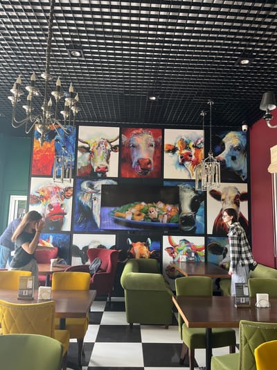 The image shows the interior of a restaurant or cafe with a black grid ceiling and a large chandelier. The walls are adorned with a grid of colorful paintings of cows. The floor is black and white checkered tile. There are several tables with wooden tops and upholstered chairs in yellow and green. In the foreground, a person wearing a black t-shirt and grey pants is walking away from the camera. Another person wearing a plaid shirt and grey pants is standing by a table. In the background, a man and a woman are standing near a wall. A security camera is visible on the wall.