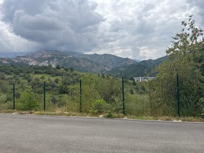 The image depicts a scenic mountainous landscape under a cloudy sky, with a paved road in the foreground. A wire fence runs horizontally across the mid-ground, separating the road from a grassy hillside dotted with trees. In the distance, a complex of buildings with white roofs is visible, nestled amongst the green slopes of the mountains. The overall scene suggests an outdoor, possibly rural or semi-rural, environment. The location is Uchterek, Uzbekistan.