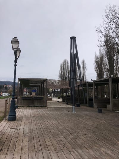 The image depicts an outdoor public area in Saverne, France, under an overcast sky. The scene is devoid of human presence or activity.

In the immediate foreground, a weathered wooden deck extends across the lower portion of the frame, showing distinct horizontal plank lines and scattered debris such as small twigs and leaves.

To the left, an ornate, dark-colored cast-iron streetlamp with two lanterns stands vertically. Adjacent to it and extending towards the center are several interconnected, open-air wooden structures, possibly kiosks or market stalls, constructed from rough timber. Some of these structures feature a layer of green material, possibly moss or netting, on their roofs. Beyond these structures, a visible rope barrier indicates proximity to a body of water or canal bank, and in the distance, residential buildings with red roofs are visible on a tree-lined hill.

In the midground, a tall, dark-colored metallic structure, composed of multiple vertical columns converging at the top into a circular opening, rises prominently. This structure appears to lean slightly. To its right, more of the wooden open-air structures are visible, with some covered by light-colored camouflage netting. The ground between these structures is a light-colored, possibly gravel or packed dirt surface, interspersed with small, dark square objects on the ground, possibly planters or bases.

The background features numerous tall, bare deciduous trees with slender, upward-reaching branches. Beyond these trees and structures, a road is visible, with a single white vehicle present. Distant hills and additional foliage complete the horizon under the uniform grey sky.