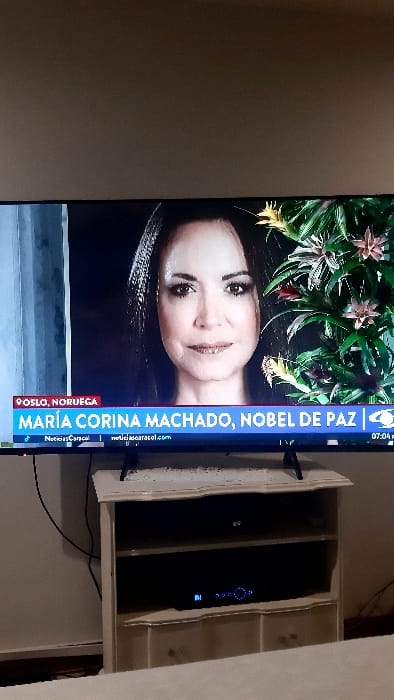 A TV screen displays an image of María Corina Machado, labeled as a Nobel Peace Prize nominee. The text on the screen says "OSLO, NORUEGA MARÍA CORINA MACHADO, NOBEL DE PAZ" and includes a URL. The woman has shoulder-length dark hair and a slight smile. The background features leafy greenery. The TV is mounted on a wall next to a white shelf unit with a black device visible inside. The time on the TV is 07:04 PM.