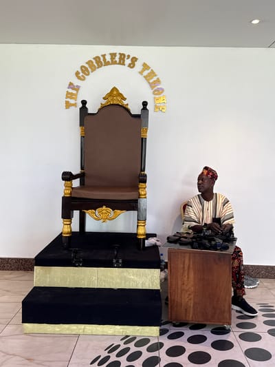 An indoor scene in Itirin, Nigeria, displays a large, ornate throne chair and a seated male figure. The throne, centrally positioned, features a dark wood frame with prominent gold accents on its armrests, legs, and back finial, and is upholstered in brown padding. It rests on a multi-tiered platform, with the top tier and two subsequent steps covered in dark fabric, each step outlined with a light gold trim. Above the throne, gold, three-dimensional letters affixed to the white wall arc to spell "THE COBBLER'S THRONE." To the right, an adult male, wearing a patterned cap, a striped shirt, and patterned trousers, sits on a partially visible chair. His hands are engaged with a collection of dark-colored sandals and shoes displayed on a small, wheeled wooden table positioned in front of him. A clear plastic water bottle is visible on the floor near the table. The floor is tiled with a light-colored surface, featuring a pattern of black circles on the right side. The background wall is plain white.