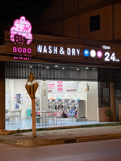 This is a night-time, eye-level, exterior shot of a commercial establishment identified by signage as "BOBO WASH & DRY" located in Baan Mae Nam, Thailand. The facade is illuminated by neon lights and features a prominent pink cartoon-like logo of a character with a bow. The main signage also reads "BOBO WASH & DRY" in illuminated letters, accompanied by icons representing Wi-Fi, a circle, and a pink circle, followed by "24 HRS". Below this, text states "wifi 24hr. cctv". The establishment appears to be a laundromat, with several washing machines visible through an open, covered area. Inside, there are tables and chairs set up. A diamond-shaped road sign with black stripes indicating a narrowing road is positioned in front of the laundromat. The surrounding environment includes a sidewalk and a paved road.