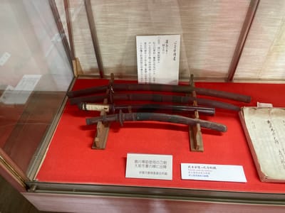 The image shows a display case in the city of Iga, Japan, featuring several antique swords. The swords are laid out on a red felt surface, supported by wooden stands. Two informational plaques are visible; one in Japanese and English, and another solely in Japanese. The Japanese and English plaque reads: "Swords used by warriors", "Swords used by warriors", and "Swords used by warriors". The other Japanese plaque reads: "Swords used by Shimakawa-so-byō\nDeployed at the Battle of Osaka in winter\nCollection of Iga City Board of Education". The display case is made of glass and metal, with a wooden base. The background appears to be a textured wall.