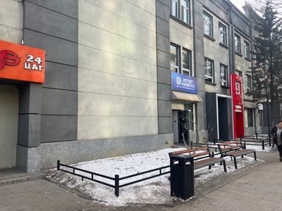 This media file captures a street scene in Ulaanbaatar, Mongolia, during what appears to be winter. The ground along the buildings is partially covered with patches of snow and slush, while the foreground features a paved sidewalk.
On the left, a building with a modern grey panel facade features an orange sign above an entrance, displaying the Cyrillic text "24 ЦАГ," which translates to "24 Hours." Adjacent to it, another building, also with a grey facade and multiple windows, has a prominent blue sign. This sign reads "ИТГЭЛТ КАПИТАЛ" (ITGELT CAPITAL) with smaller text below: "БАНК БУС САНХҮҮГИЙН БАЙГУУЛЛАГА," meaning "NON-BANK FINANCIAL INSTITUTION." Below this sign is an entrance.
Further to the right, a tall, vertical red structure with a stylized logo (partially obscured) marks another business entrance. In front of these buildings, a pedestrian area features three benches made of wood and black metal, along with a matching black metal trash can. A low black metal fence separates the snowy ground from the paved sidewalk. Bare trees and a single evergreen tree stand along the building line. A person, partially visible on the far right, is walking on the sidewalk. The sky above appears overcast, indicating daytime.