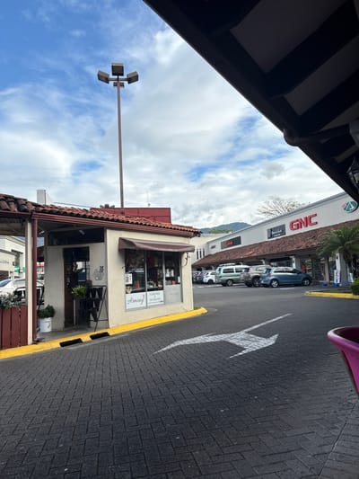 The image shows a street corner in Pozos, Costa Rica, at the coordinates 9.951 latitude and -84.194 longitude. The scene is dominated by a brick-paved street with a white arrow painted in the middle, leading into a parking lot on the right. The parking lot is lined with a row of shops with white facades. The most prominent shop on the right has a red "GNC" sign. There are several parked cars in the lot, including a white van, a grey car, and a silver car. The image captures a view from the edge of the street, looking towards the parking lot. The building on the left is a small shop with a brown awning and a sign reading "Armony." There is a streetlight with four lights in the middle of the image. The sky is blue with white clouds. The background is a mountain range that is visible through the buildings.