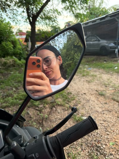 The image displays a female individual taking a selfie, reflected in the left rearview mirror of a motorcycle, located in Benoa, Indonesia. The reflection shows a woman with dark hair, wearing round eyeglasses, a black choker necklace, and a white t-shirt. She holds an orange-cased smartphone with a triple-lens camera system in her right hand, which has visible finger tattoos. Below the mirror, the black handlebar grip and brake lever of the motorcycle are visible. The external environment, seen directly beneath the mirror and reflected within it, consists of unpaved ground with sparse green vegetation. In the background reflected by the mirror, a grey SUV is parked under a simple, open-sided shelter with a corrugated roof, surrounded by green foliage and distant residential structures under a light sky.