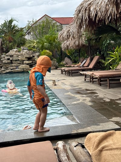A young child wearing an orange sunsuit and hat stands at the edge of a swimming pool, looking down into the water. In the pool, a person wearing a sun hat and a pink float is visible. The poolside is paved with stone and lined with lounge chairs under a thatched roof structure. In the background, there is a stone waterfall feature and lush tropical vegetation, as well as a building with a red tile roof. The scene appears to be a resort or hotel in Nam Hoa Lư Ward, Vietnam.