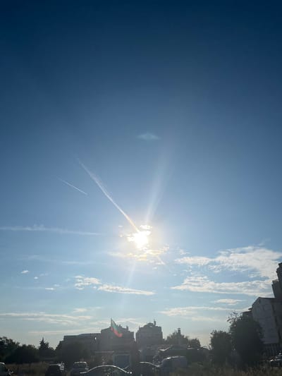 The image captures a clear, bright sky over the city of Nesebar, Bulgaria. The sun is positioned in the upper-middle section of the frame, casting strong rays and creating a lens flare effect. A faint airplane contrail stretches diagonally across the sky from the upper left to the lower right, with another fainter contrail to its left. Below the sky, the urban landscape of Nesebar is visible. Several buildings of varying architectural styles are present, some appearing modern and others with more traditional elements, including domed roofs. A Bulgarian flag, colored white, green, and red, is prominently displayed on a flagpole against the backdrop of buildings. In the foreground, the image shows a street scene with parked cars, some covered with tarps, and dense green foliage, suggesting an outdoor environment within the city. The overall scene depicts a sunny day in the Bulgarian coastal city.