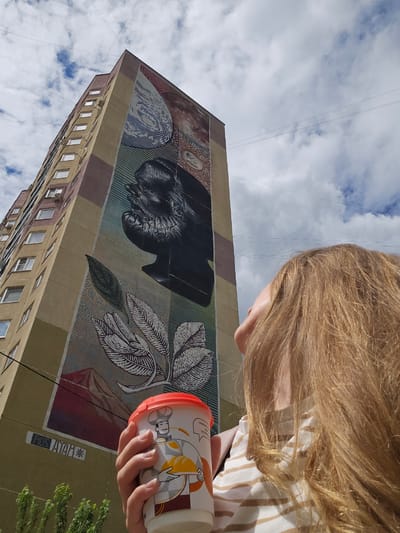 In Odintsovo, Russia, a young woman with blonde hair holds a disposable cup with a chef depicted on it. She is standing in front of a tall apartment building adorned with a large mural. The mural features abstract designs, a close-up of a person's face in profile, and botanical illustrations of leaves. The building has multiple windows and air conditioning units. The sky above is partly cloudy, with visible power lines crisscrossing the frame. A street sign with the Cyrillic text "PIOS LATAM" and a star is visible near the bottom of the building.