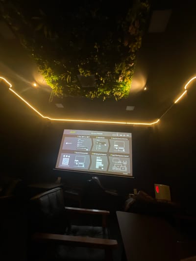 This is a medium shot of a projector screen displaying setup instructions for an Acer projector. The screen is illuminated by the projector's light, with a portion of a wall featuring a vertical garden visible to the left. Yellow LED strip lighting outlines a section of the wall and ceiling. To the right of the screen, there are glimpses of dark furniture, possibly chairs and a table, suggesting an indoor seating area. A small white device with a red light is mounted on the wall above the furniture. The overall scene is dimly lit, creating a dark ambiance, and the orientation of the image suggests it was taken in portrait mode. The visible text on the screen includes "Acer," "ID: Acer-F70A4A8E," "1 Press +K or +P," "2 Select Device," "3 Start Your Screen Mirroring," "Windows 10/11," "1 Search & Connect WiFi," "2 WiFi Setup For Projector," "3 Start The Projection Control," and a serial number "21973423." The low lighting and the projector setup indicate the image was likely taken at night or in a dark room.