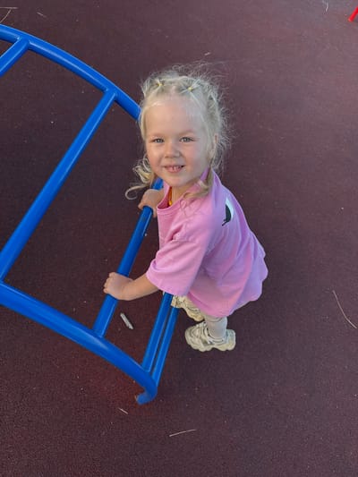 A young girl with blonde hair tied in pigtails is on a playground. She is wearing a pink t-shirt with a black graphic and grey pants, along with white sneakers. The girl is lying on a blue monkey bar structure, with her hands gripping the bars. She is smiling at the camera, appearing happy and engaged. The playground surface is a dark, textured material, possibly rubberized, common in modern playgrounds. The background is a blur of the same dark surface, suggesting an outdoor setting. The image appears to be taken during the daytime, with natural light illuminating the scene. The girl's pose and expression convey a sense of playfulness and enjoyment of the playground equipment. There is no visible text in the image.