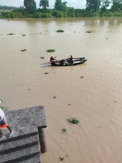 This image depicts a view of the Nun River in Yenagoa, Nigeria. The river is murky brown and covered with scattered water hyacinths. In the middle of the river, a wooden canoe with two occupants is visible. One person is seated towards the back, steering with a long oar, and the other is seated in front. The canoe contains fishing nets and other supplies. On the left side of the image, a concrete structure with rough texture serves as a dock or embankment. A person wearing an orange and white outfit with grey flip-flops is standing on this structure. The background features a dense line of green vegetation and trees along the riverbank. The sky is overcast, with soft clouds.