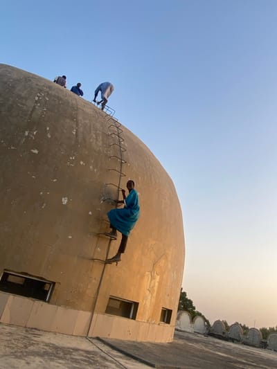 This image depicts a large, light-brown, concrete dome-shaped building in Barako, Nigeria, under a clear, pale blue sky. A metal ladder, consisting of vertical bars and horizontal rungs, is affixed to the curved surface of the dome, extending from its base towards the apex.
Four adult males are visible on the dome. In the mid-ground, a man wearing a blue robe (jalabiya), dark trousers, and sandals is actively climbing the ladder, ascending with his right foot on a rung and left leg bent. He is turned slightly towards the viewer, exhibiting a smile. Above him, closer to the dome's crest, three other men are positioned. One man in a light-colored robe is seen from behind, bent over, appearing to secure or interact with the upper section of the ladder or the dome's surface. Two other men are partially visible further up the dome, one in a dark garment and another in a light blue garment, both observing or positioned near the dome's highest point.
The dome's surface shows signs of wear, including visible cracks and areas of discoloration. Below the ladder, on the lower section of the dome, multiple rectangular window openings are present. The immediate foreground shows a flat concrete rooftop area with a decorative, curved parapet wall extending into the distance on the right. Trees are visible on the horizon line beyond the parapet. The lighting suggests late afternoon or early morning.