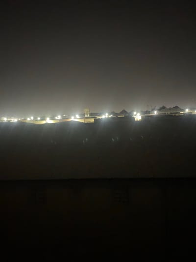 The image depicts a nighttime view of an urban landscape in Fagge A, Nigeria. The foreground is dominated by a dark, textured surface, possibly a wall or ground, with subtle hints of reflected light. Beyond this, a row of buildings stretches across the mid-ground, illuminated by numerous bright, scattered lights. These lights appear to be streetlights or lights from within the structures, creating a hazy glow in the dimly lit environment. In the far distance, the silhouette of a water tower is visible, along with the faint outlines of more buildings and possibly some vegetation against the dark, overcast sky. The overall impression is one of a city at night, with artificial lighting cutting through the darkness and illuminating the built environment.