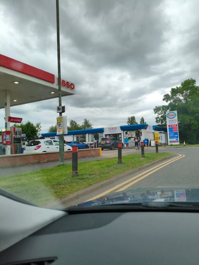 An eye-level, medium shot captures an exterior scene at a Tesco Express petrol station on a cloudy day. The Esso branding is prominent, with a large red and white sign featuring the Esso logo and "Tesco express" text, alongside fuel prices. Several cars are visible, including a white hatchback at a pump, a dark SUV, and a blue sports car. Two individuals, both appearing to be male, are near the entrance of the Tesco Express store. The foreground shows the interior of a vehicle, with the dashboard and windscreen visible, reflecting some of the scene. The environment includes a paved road with yellow lines, a grassy area, and trees in the background. The location is identified as Wychavon, United Kingdom.