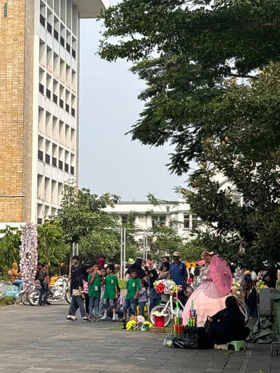 The image depicts a scene in a public outdoor area with a tall, beige building with many windows on the left. In the foreground and midground, there is a gathering of people, many of whom appear to be children, some wearing green t-shirts. There are also adults present. Several objects are visible, including bicycles, motorcycles, colorful flowers arranged as decorations, and umbrellas. A person dressed in a large pink gown and hat, holding a pink umbrella, is a prominent figure in the center. In the background, more trees and buildings can be seen. The overall atmosphere suggests a public event or gathering, possibly a market or festival. While the specific location within West Jakarta, Indonesia, is not explicitly stated in the image, the presence of a large building and an outdoor gathering area is consistent with urban environments in that region.