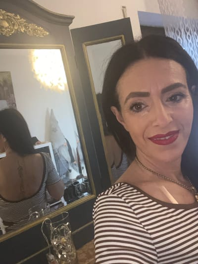 A woman with dark hair and bright red lipstick poses in front of a large ornate mirror. The mirror reflects the same woman holding a phone, presumably taking the photograph. The background suggests a bedroom setting with a feather boa and decorative lights visible. The woman is wearing a striped top. The location is not specified as Konz, Germany.