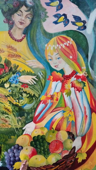 The image is a painting depicting two women and a basket of fruit. The woman on the left has dark hair adorned with flowers and wears a yellow dress with a beaded necklace. Her hand gestures towards a cluster of green foliage with blue and red flowers. The woman on the right has blonde hair styled with white accents and wears a colorful dress decorated with autumn leaves. She is looking towards the viewer and reaching into a basket filled with various fruits including apples, grapes, and other round fruits. The background of the painting features abstract blue and green elements and three stylized birds in flight. A "220 V" label is visible on the right side of the painting. The painting is an artistic representation and not a photograph of a specific location. Therefore, it is not possible to determine its location in the city of Kryvyi Rih, Ukraine based solely on the image.
